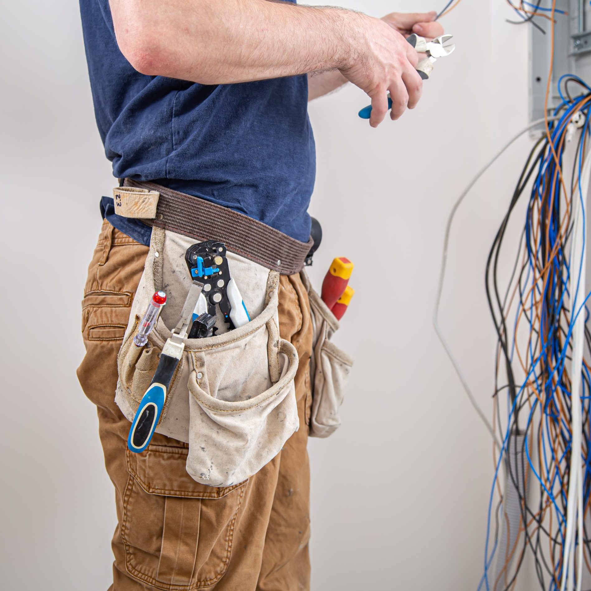 Electrician builder examines the cable connection in the electrical line in switchboard indoor. Electrician builder at work, examines the cable connection in the electrical line in the fuselage of an industrial switchboard. Professional in overalls with an electrician's tool.